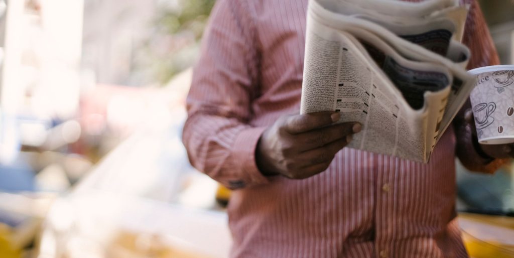 Crop unrecognizable male reading newspaper and holding paper cup with hot beverage while standing on city street — Cheap Cryp